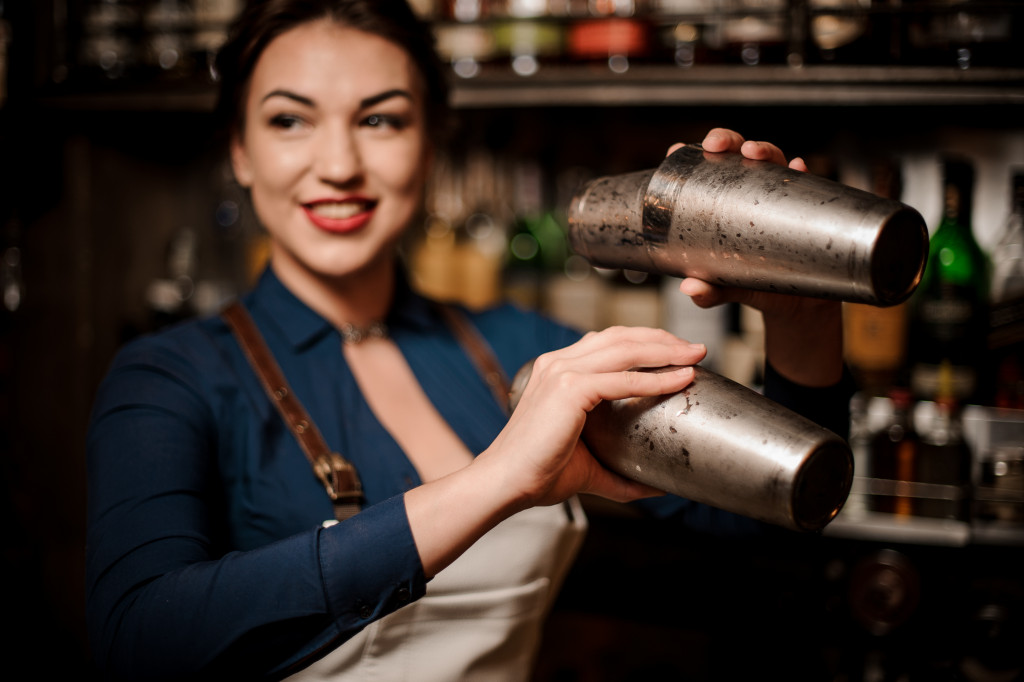 Bartender in the white apron holding in her hands two steel cocktail shakers at the bar counter